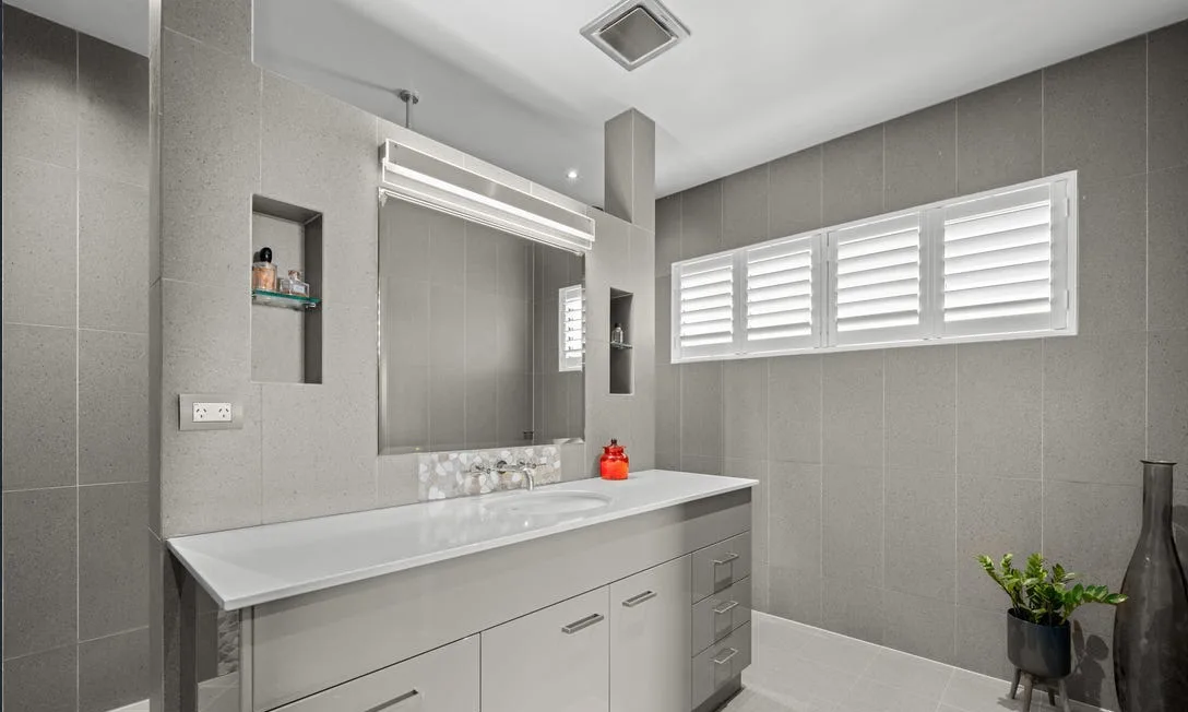 Grey-tiled bathroom with a long vanity, plantation shutters and a floor-standing vase