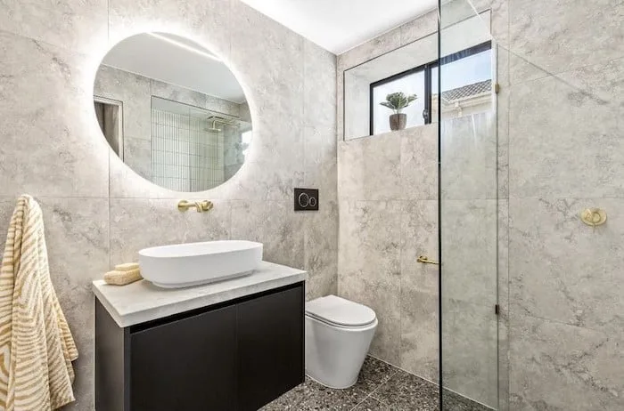 Ensuite with a round backlit mirror, gold tapware and a light stone vanity